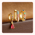 Three gold huggie earrings with rainbow stones and a red teardrop charm