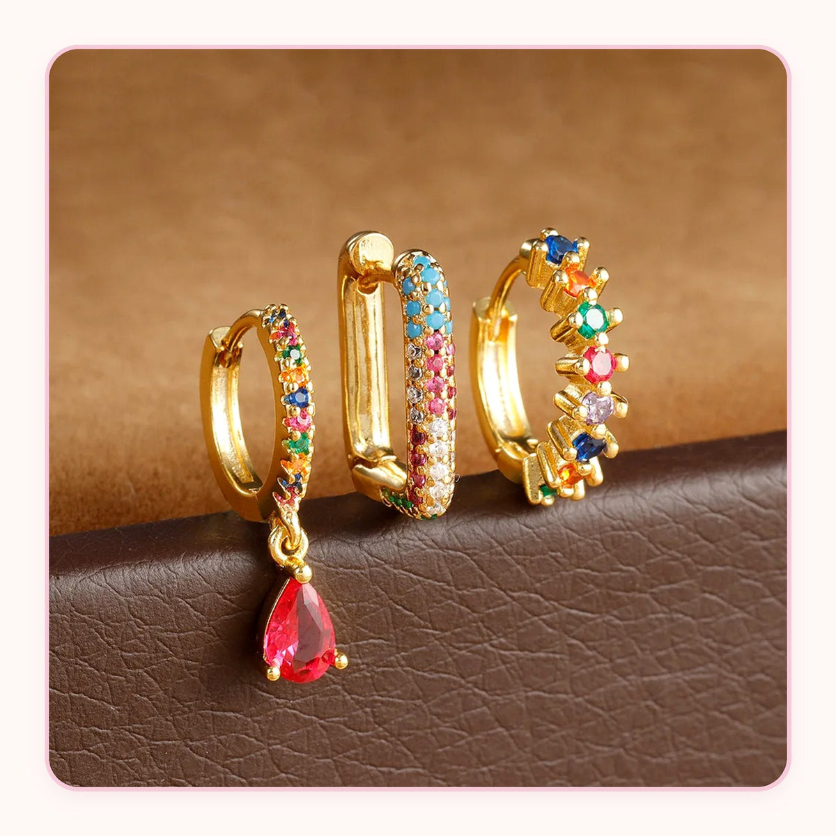 Three gold huggie earrings with rainbow stones and a red teardrop charm