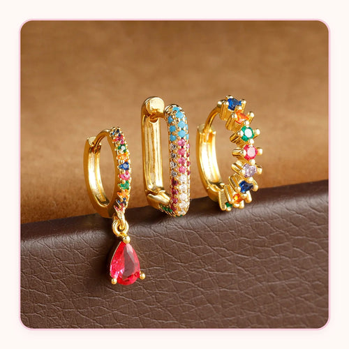 Three gold huggie earrings with rainbow stones and a red teardrop charm